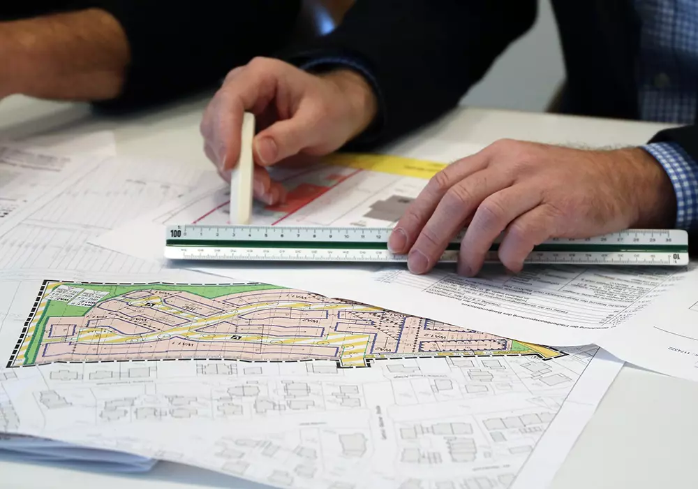 Zwei Hände arbeiten mit Maßstab und Stift an einem farbigen Bebauungsplan und technischen Zeichnungen auf einem Schreibtisch.