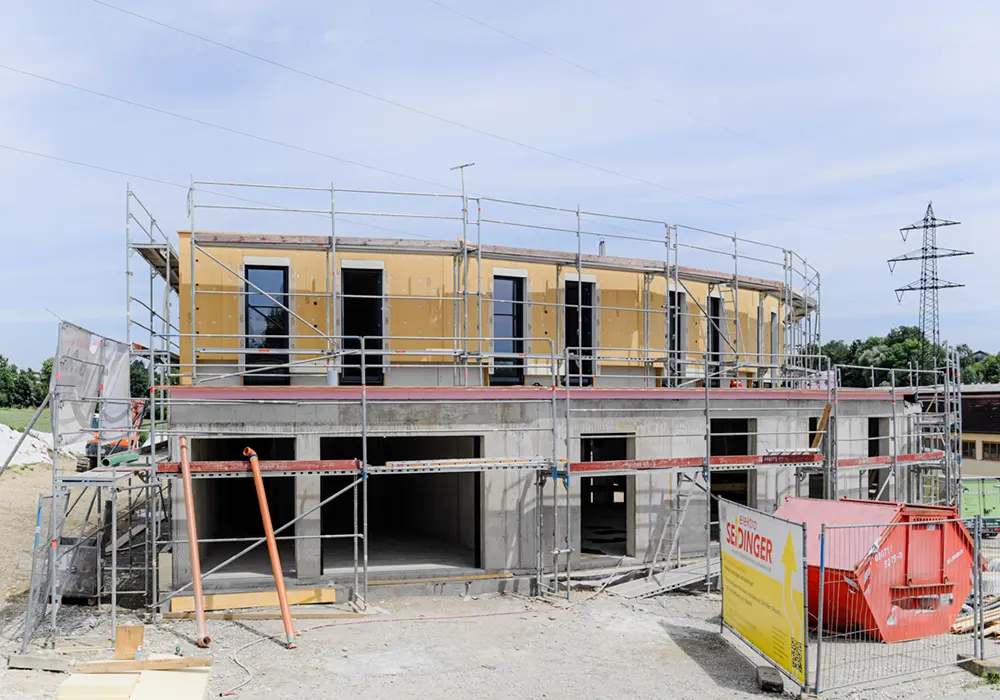 Zweistöckiges Gebäude im Rohbau mit Gerüst, gelber Container und Strommast im Hintergrund.