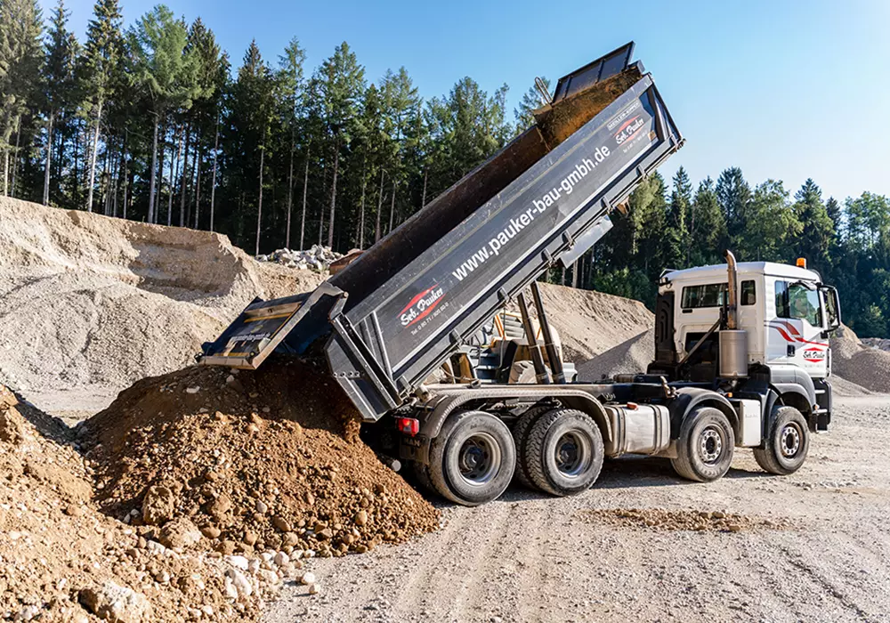Kipplaster der Firma Pauker-Bau entlädt Erdmaterial auf einem Baustoffplatz mit Kies- und Sandhügeln; im Hintergrund dichter Nadelwald bei sonnigem Wetter.