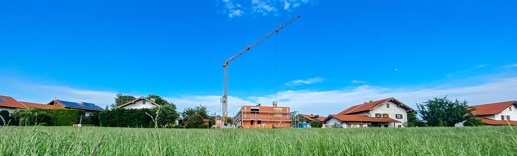 „Neubau mit Baukran auf freiem Feld, eingerahmt von Wohnhäusern mit roten Dächern unter strahlend blauem Himmel.