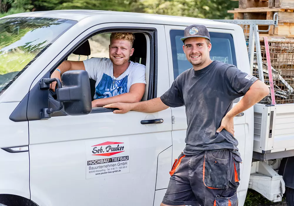 Zwei junge Bauarbeiter lachen in Richtung Kamera, einer sitzt im Firmenfahrzeug, der andere steht daneben.