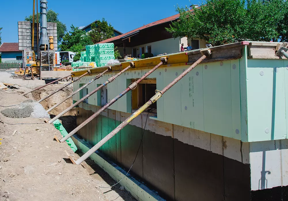 Gedämmter Kellerrohbau mit Abstützungen und offener Baugrube auf einer Baustelle bei Sonnenschein.