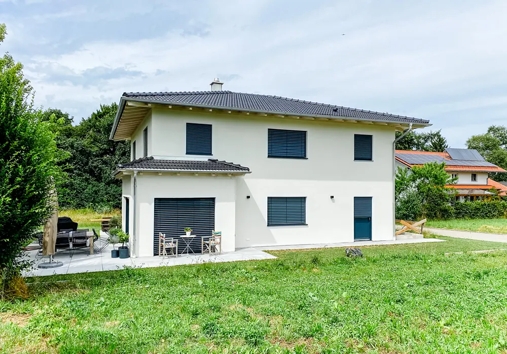 Modernes weißes Wohnhaus mit dunklem Walmdach, geschlossenen Rollläden und möblierter Terrasse im Grünen.