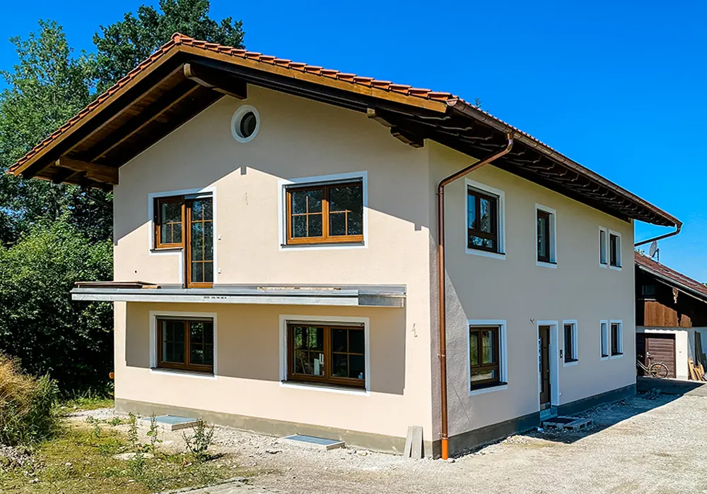 Zweigeschossiges Haus mit hellem Putz, Holzfenstern und rotem Ziegeldach, umgeben von Kiesfläche und Bäumen.