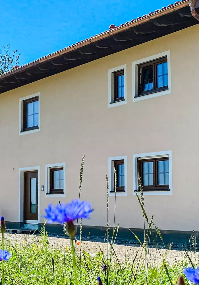 Wohnhaus mit beigem Putz, braunen Fensterrahmen und Wildblumen im Vordergrund.