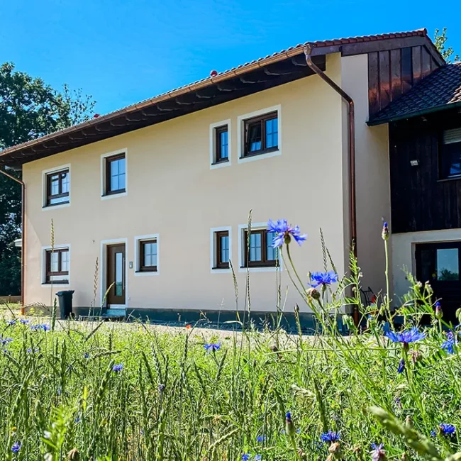 Zweifarbiges Wohnhaus mit Wildblumenwiese im Vordergrund, bei sonnigem Wetter vor Bäumen fotografiert.