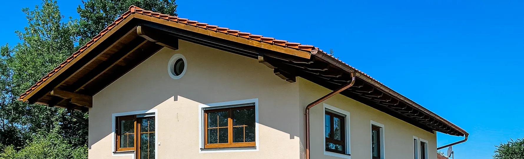 Giebel eines Hauses mit roten Dachziegeln, Holzfenstern und rundem Oberlicht vor blauem Himmel.