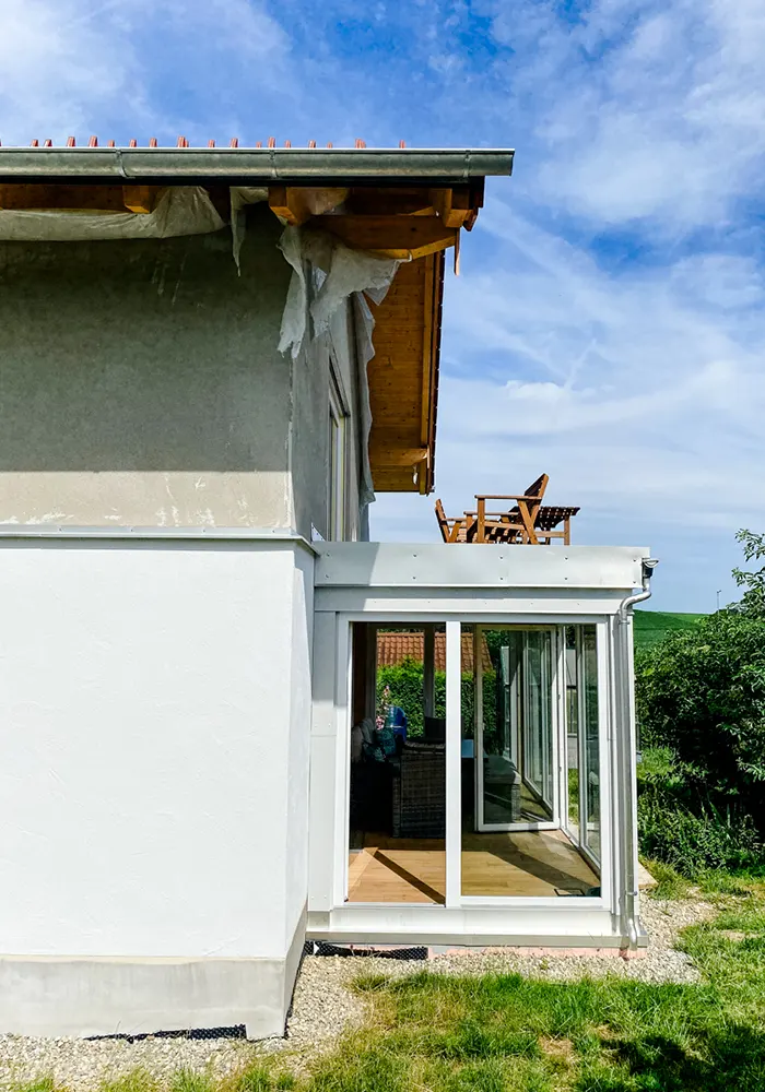 Haus mit Wintergarten im Erdgeschoss und Terrasse mit Holzmöbeln im Obergeschoss, Blick von der Seite.