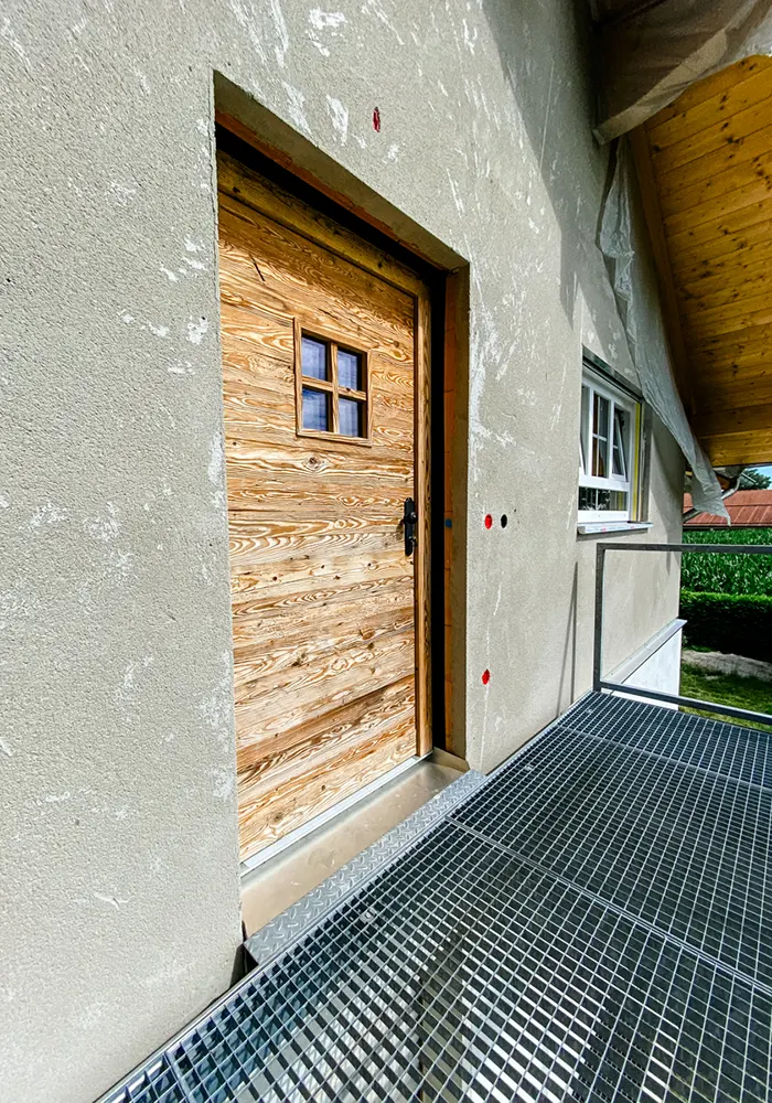 Holztür mit kleinem Fenster auf Metallbalkon an grauer Hausfassade, daneben weißes Fenster und Geländer.