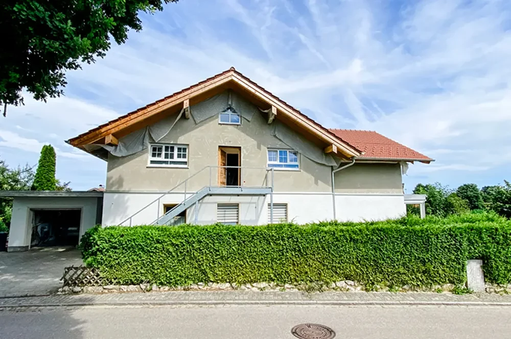 Zweistöckiges Haus mit rotem Satteldach, Außentreppe und umlaufender grüner Hecke vor blauem Himmel.