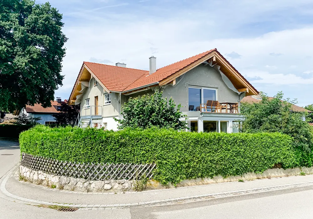 Eckhaus mit rotem Satteldach, Balkon und grüner Hecke, gelegen an einer gepflaster Straße.