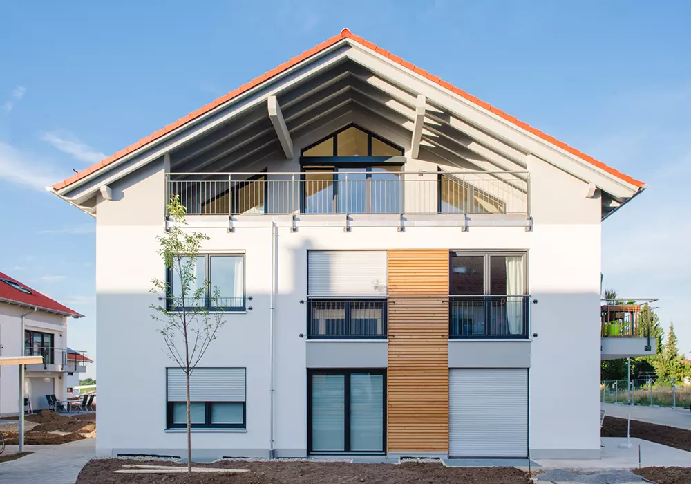 Neubau eines weißen Mehrfamilienhauses mit Balkon, Holzfassadenelementen und rotem Satteldach bei blauem Himmel.