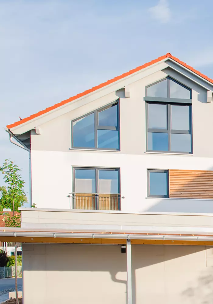 Oberer Gebäudeteil eines modernen Hauses mit großen Fenstern, rotem Satteldach und Holzverkleidung bei Sonnenschein.