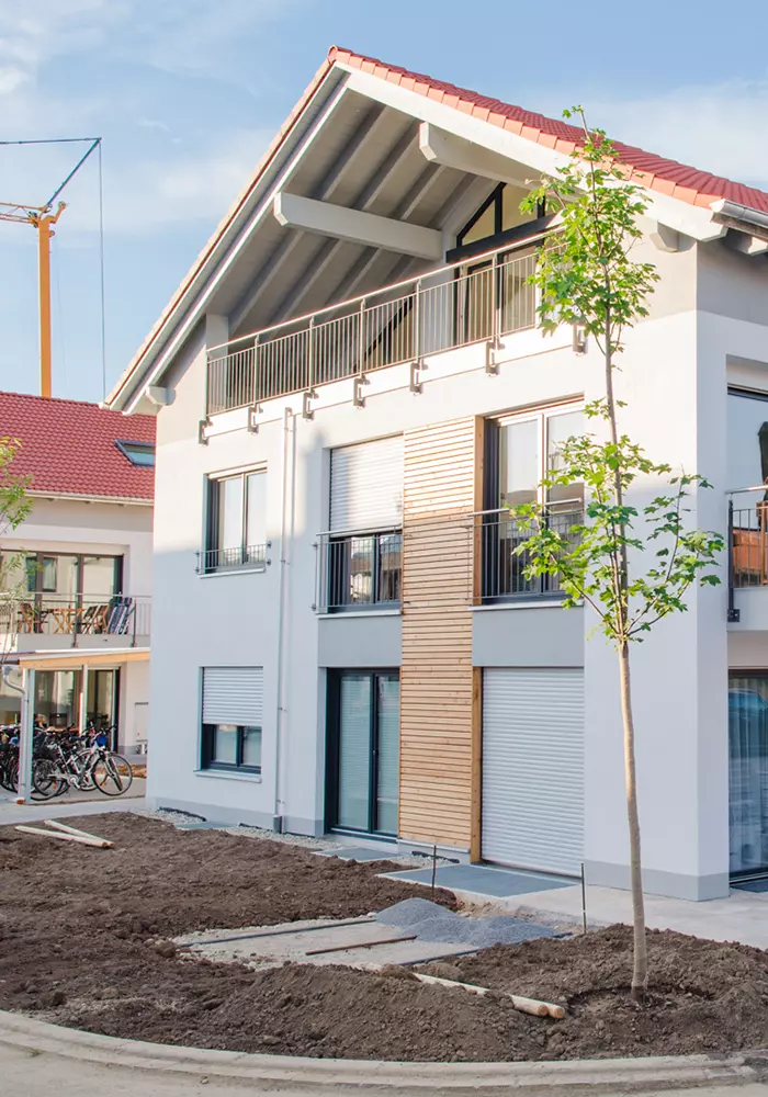 Moderne Wohngebäude in einem Neubaugebiet mit weißer Fassade, großen Fenstern und Holzelementen an der Fassade; der Außenbereich ist noch im Bau mit unbepflanzter Erde und Pflastersteinen.