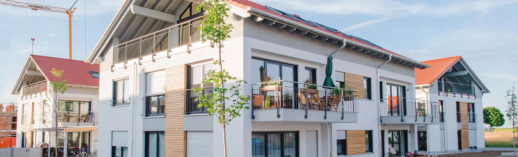 Mehrfamilienhaus mit roten Dächern, Balkonen und jungen Bäumen, Neubaugebiet mit noch unbepflanzten Flächen.