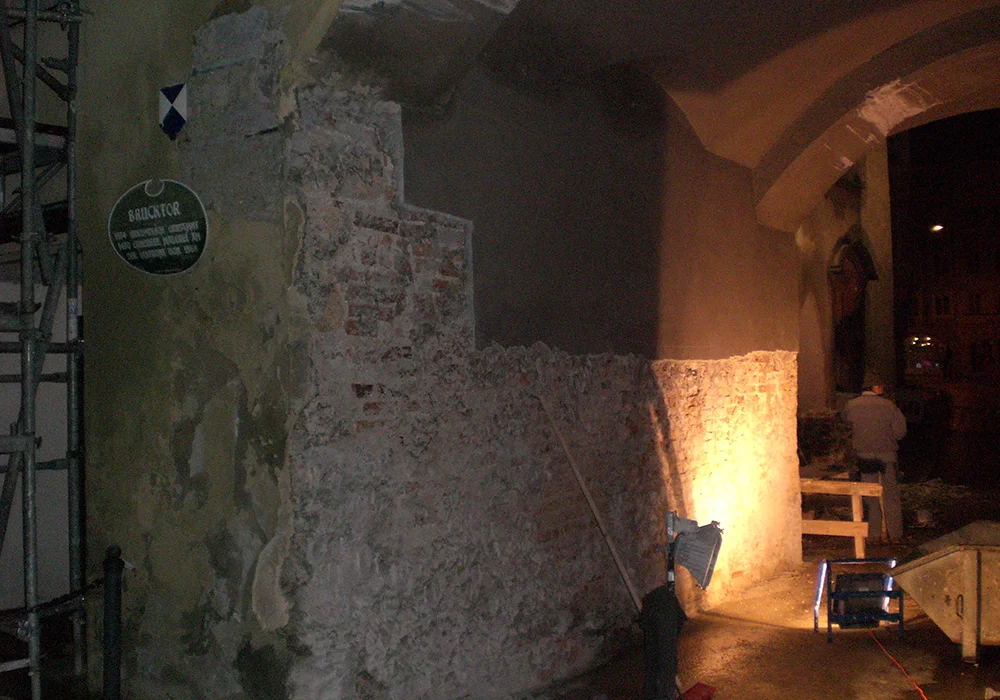 Teilweise freigelegte, verwitterte Ziegelmauer eines historischen Gebäudes mit einem grünen Hinweisschild ‚Brucktor‘ und Baugerüst im Hintergrund.
