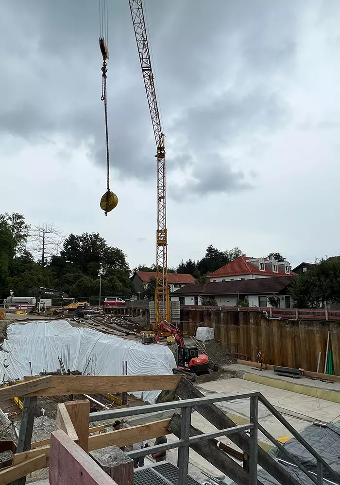 Baustelle mit Kran, Bagger und gesicherter Baugrube, abgedeckt mit Folien, vor Wohnhäusern und bewölktem Himmel.