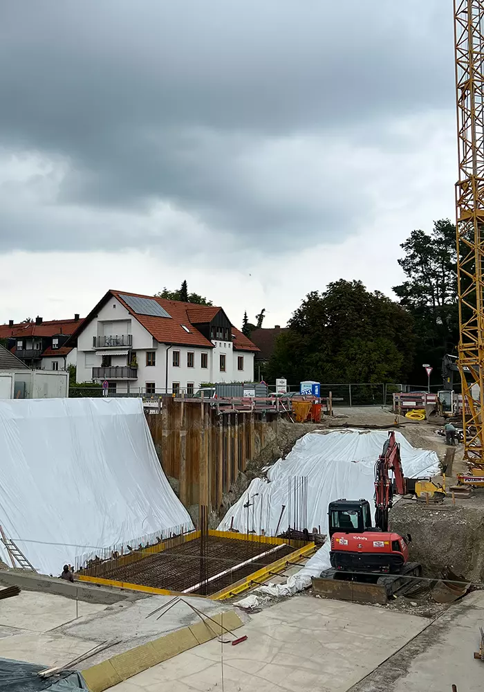 Tiefbaugrube mit Bagger und Baukran vor Wohnhäusern, gesichert mit Folien und Spundwänden unter bewölktem Himmel.