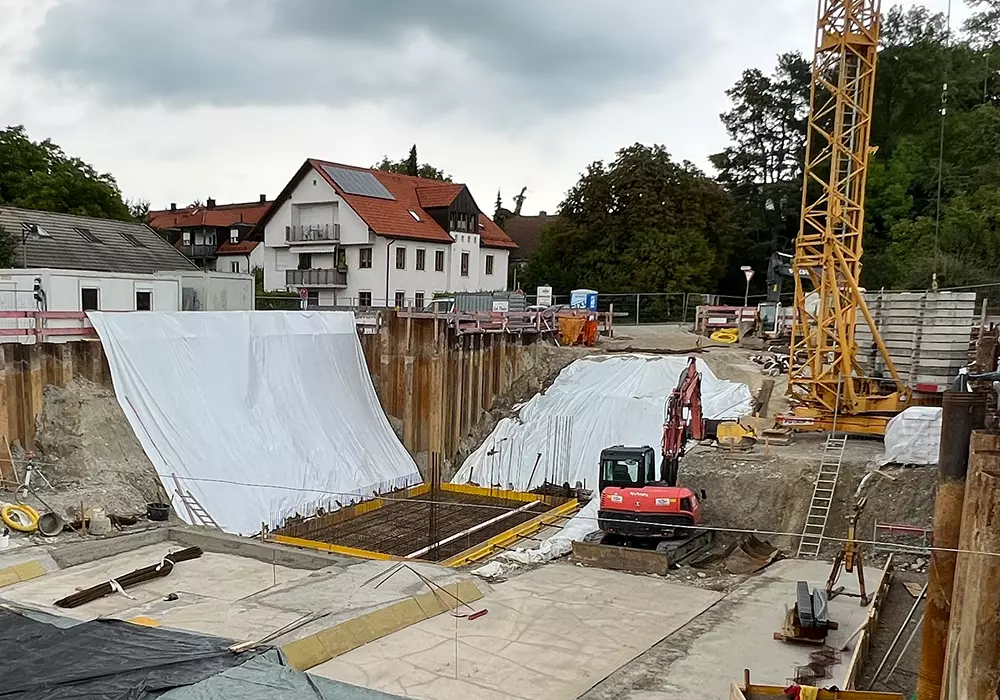 „Große Baugrube mit Folienabdeckung, Bagger, Kran und Stahlbewehrung vor Wohnhäusern und bewölktem Himmel.