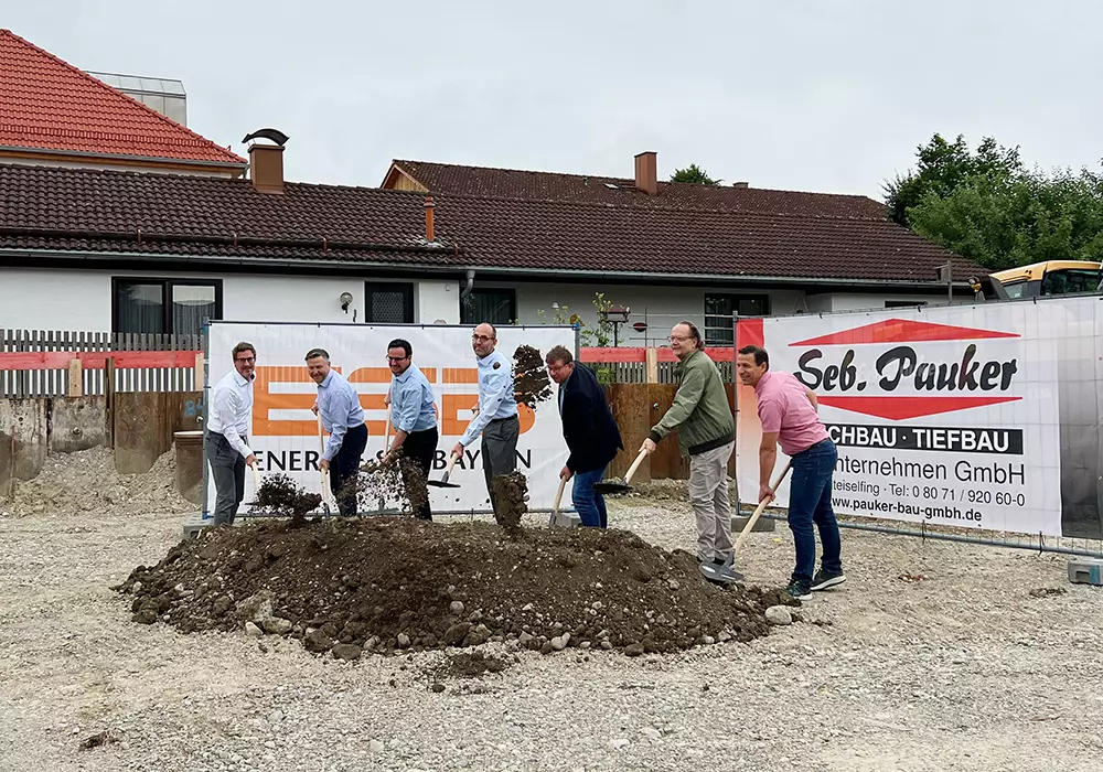Sieben Personen beim Spatenstich auf einer Baustelle vor zwei Werbebannern und Wohnhäusern.