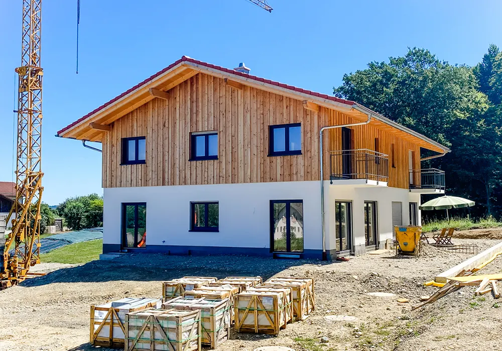 Neubau mit Holzfassade im Obergeschoss, großen Fenstern und Balkonen, Baustellenmaterial im Vordergrund.
