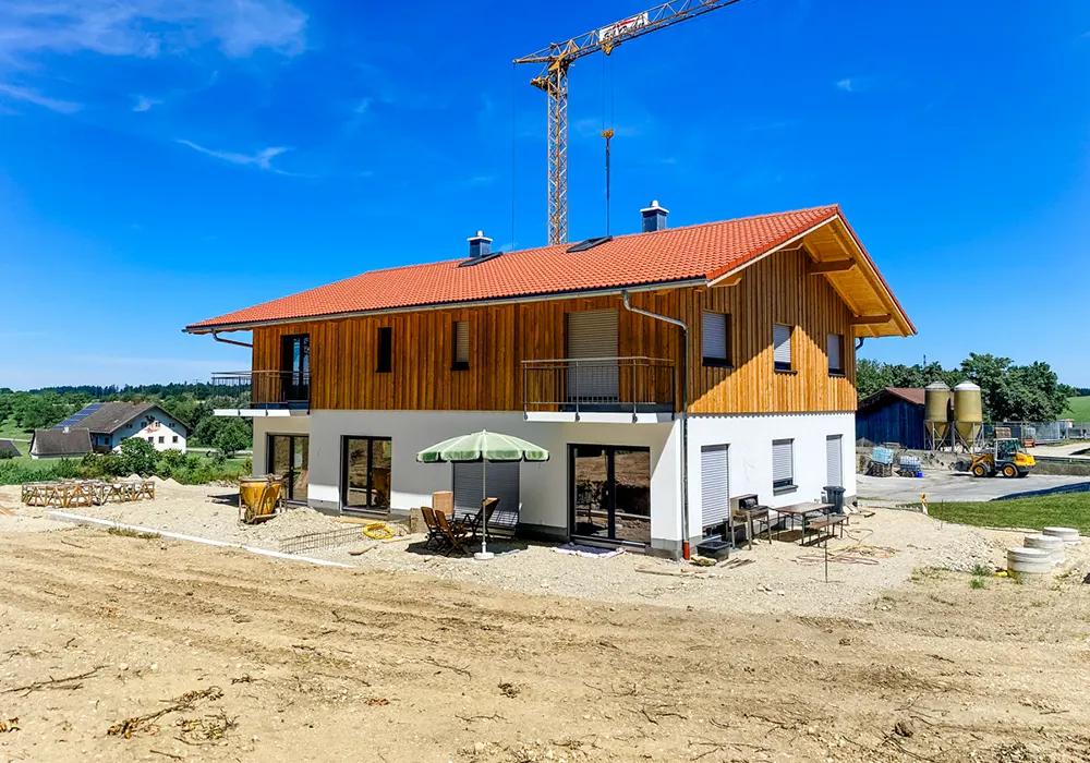 Zweigeschossiges Haus mit Holzfassade, roten Ziegeldach, Balkon und Baustelle im ländlichen Umfeld.