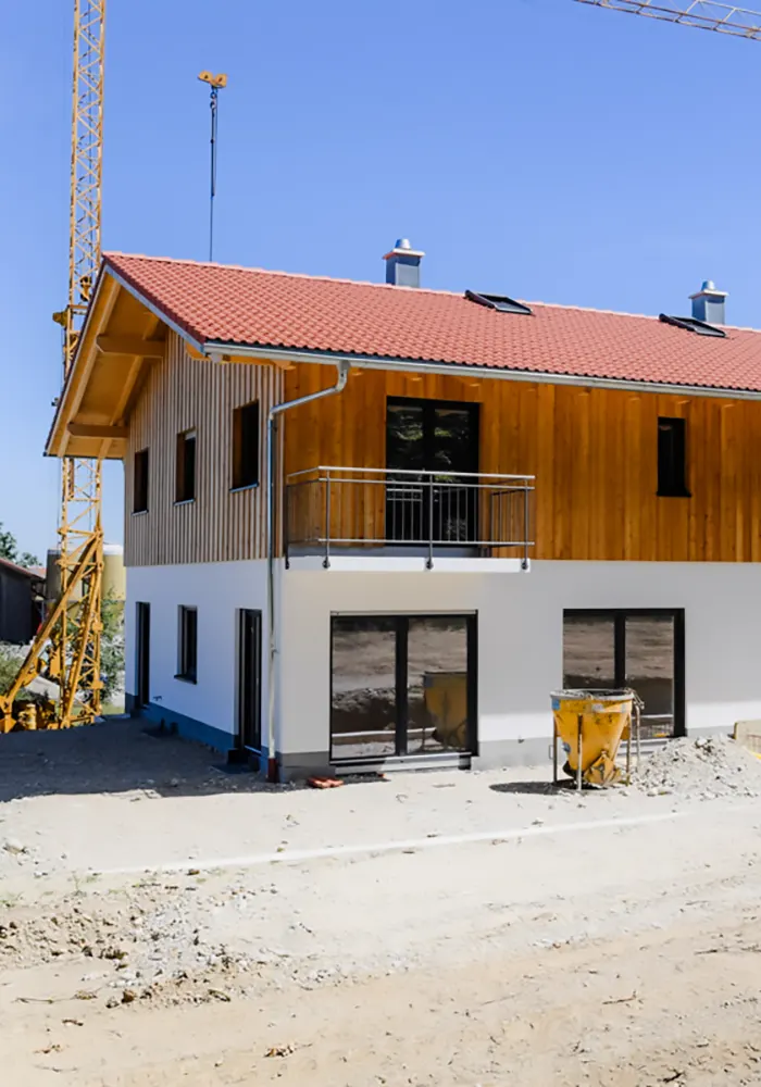Neubau mit Holzfassade, Balkon und roten Dachziegeln, Baukran und Schüttmulde im Vordergrund.