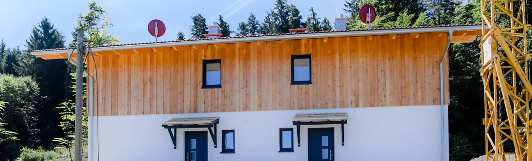 pauker-bau-gmbh-referenzen-neubau-dh-pfaffing-header Doppelhaushälfte mit Holzfassade im Obergeschoss, dunklen Fenstern und Haustüren, vor Waldkulisse.
