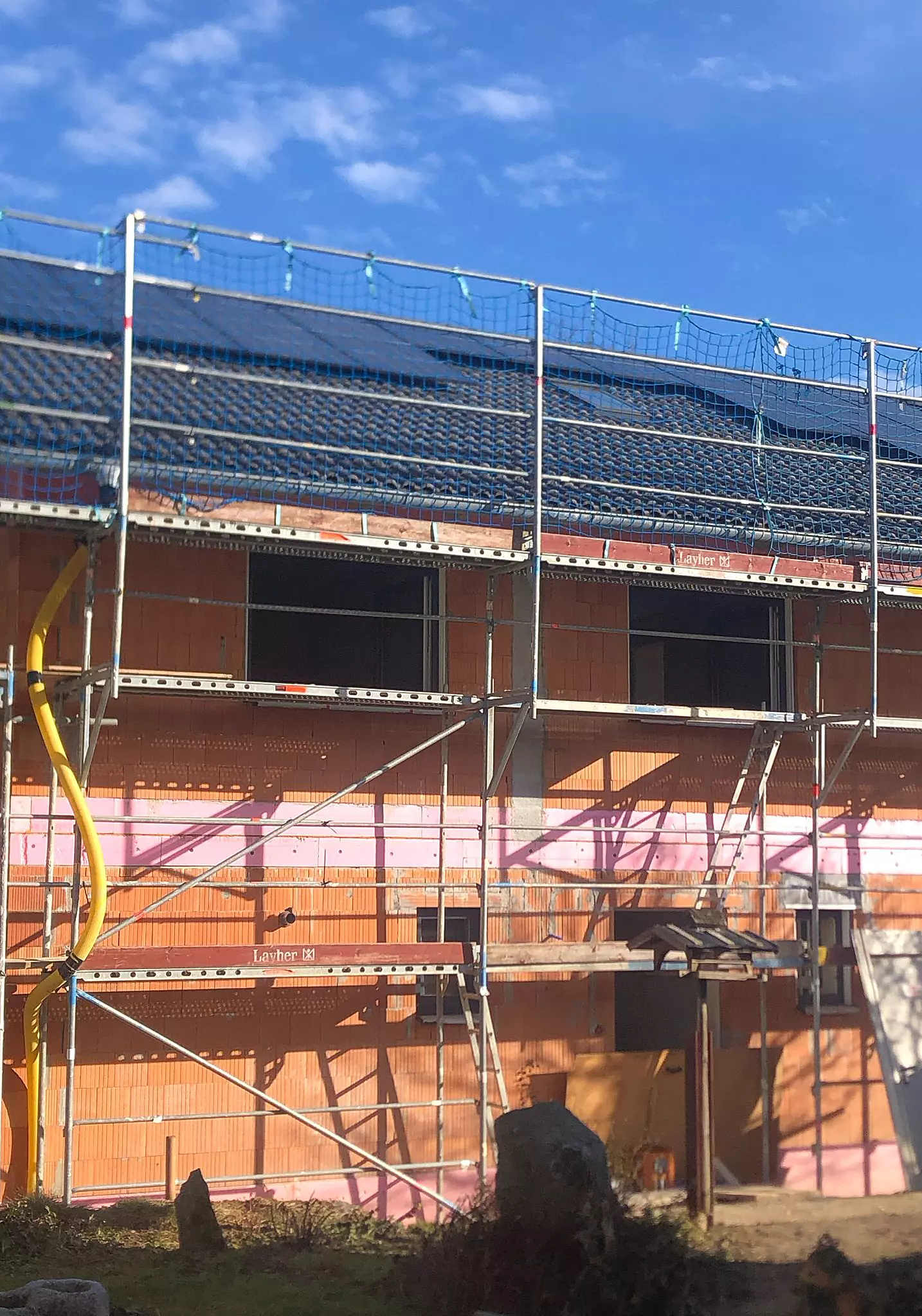 Zweistöckiger Rohbau mit Gerüst und gelbem Bauschutt-Rohr, Ziegelmauerwerk und Photovoltaikanlage auf dem Dach.