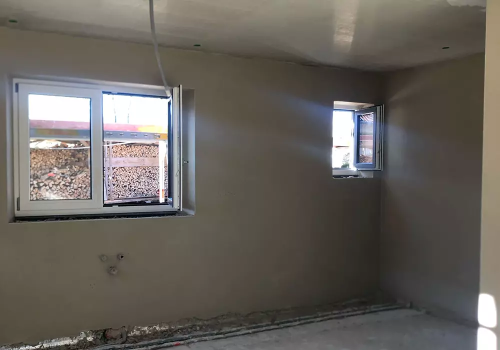 Innenaufnahme eines im Bau befindlichen Raums mit zwei geöffneten Fenstern, unverputzten Wänden und sichtbaren Leitungen am Boden.