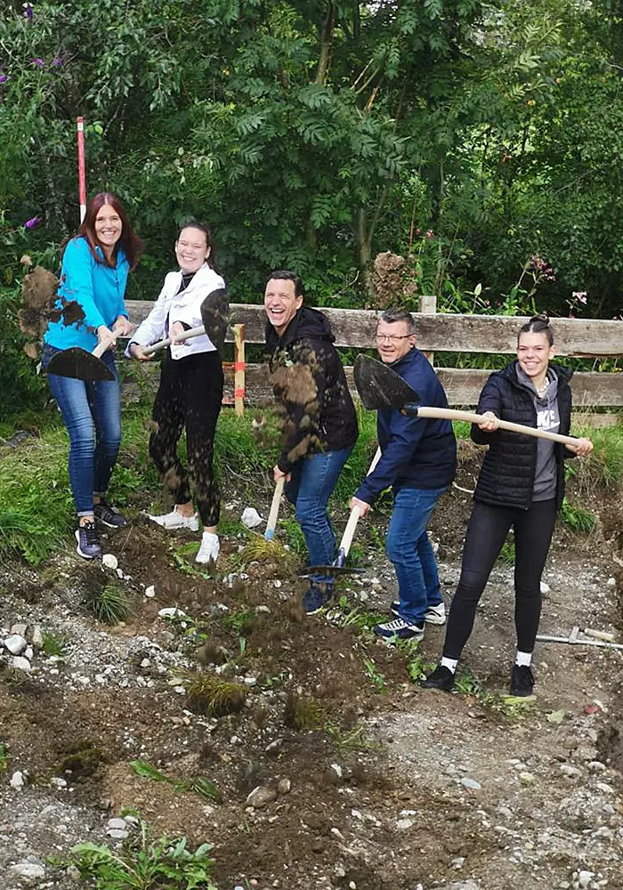Fünf lachende Personen mit Schaufeln beim symbolischen Spatenstich auf einer begrünten Baustelle.