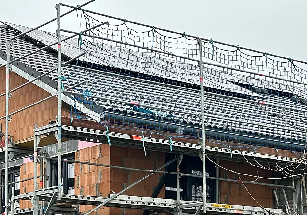 Haus im Rohbau mit Ziegeldach und installierten Solarpanels, eingerüstet mit Baugerüst und Sicherungsnetz.