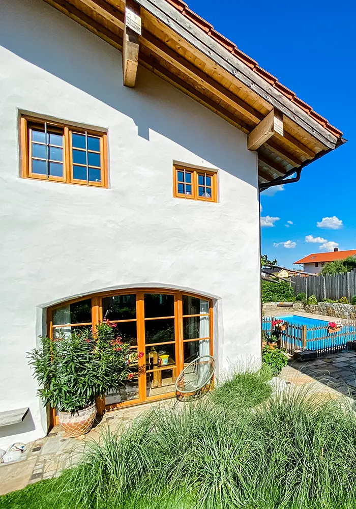 Weiße Hausfassade mit Holzfenstern, davor hohe Gräser, ein Blumentopf und Blick auf eingezäunten Poolbereich.