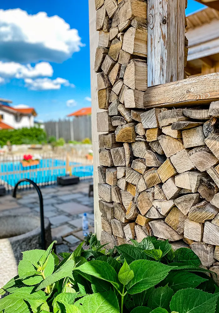 Gestapeltes Brennholz an einer Holzwand, im Vordergrund grüne Blätter, im Hintergrund Hof mit Pool und blauer Himmel.