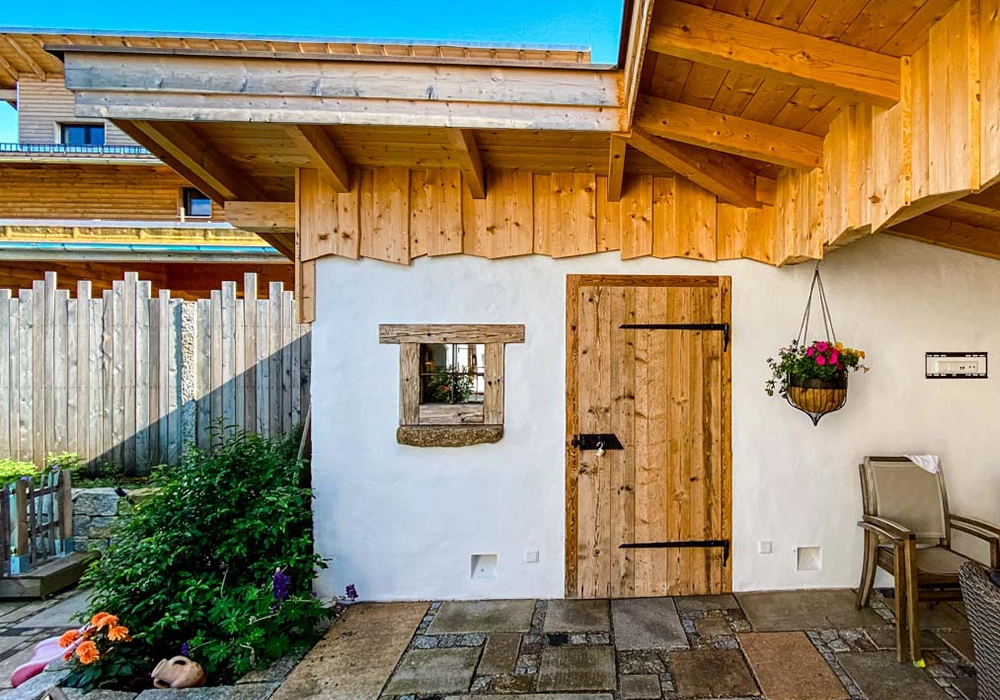 Holzverkleideter Eingangsbereich mit Holztür, kleinem Fenster, Hängeblumen und Gartenmöbeln.