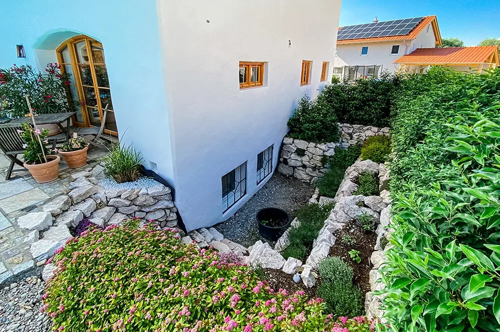 Terrassierter Garten mit Steinmauern und Blumenbeeten an weißem Haus, daneben Sitzbereich mit Blumentöpfen.