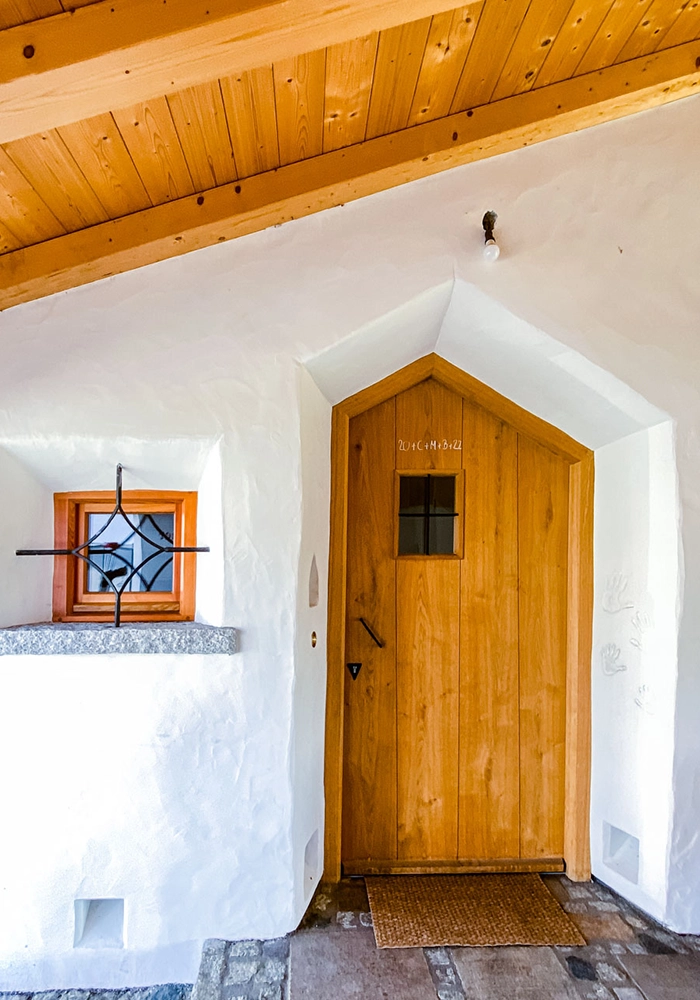 Überdachter Hauseingang mit spitzer Holztür, kleinem vergitterten Fenster und Steinboden.