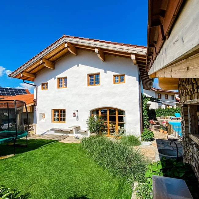 Gepflegter Garten mit Trampolin und Sitzbank vor einem Haus mit Holzfenstern, daneben Pool und Holzlager.