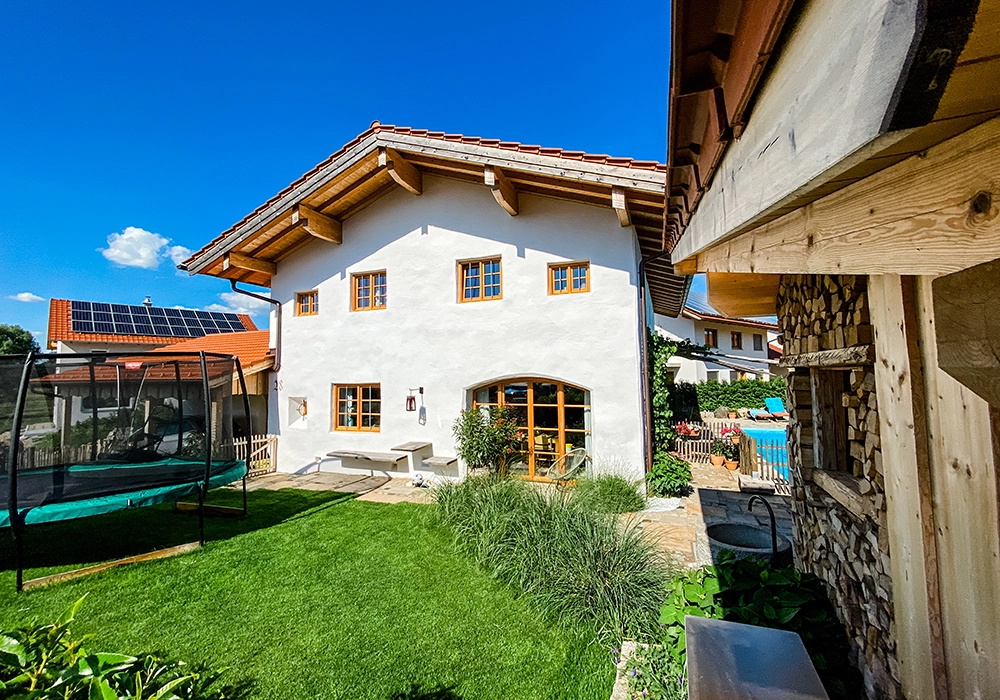 Gepflegter Garten mit Trampolin und Sitzbank vor einem Haus mit Holzfenstern, daneben Pool und Holzlager.