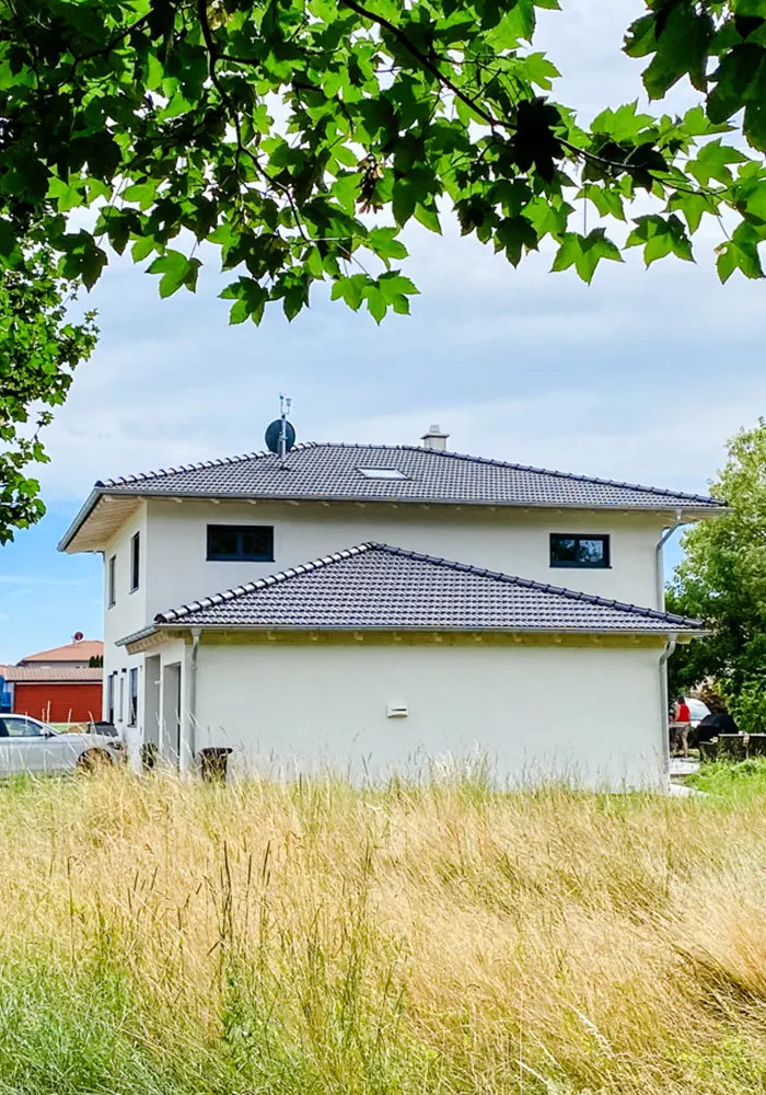 Zweistöckiges Einfamilienhaus mit dunklem Walmdach hinter einer Wiese mit hohem Gras und Bäumen.
