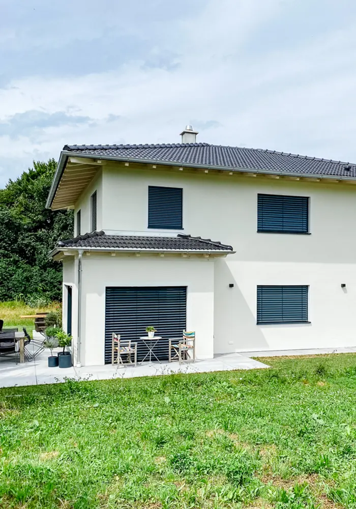 Modernes weißes Haus mit Flachdach, geschlossenen Rollläden und kleiner Terrasse mit Sitzmöbeln.