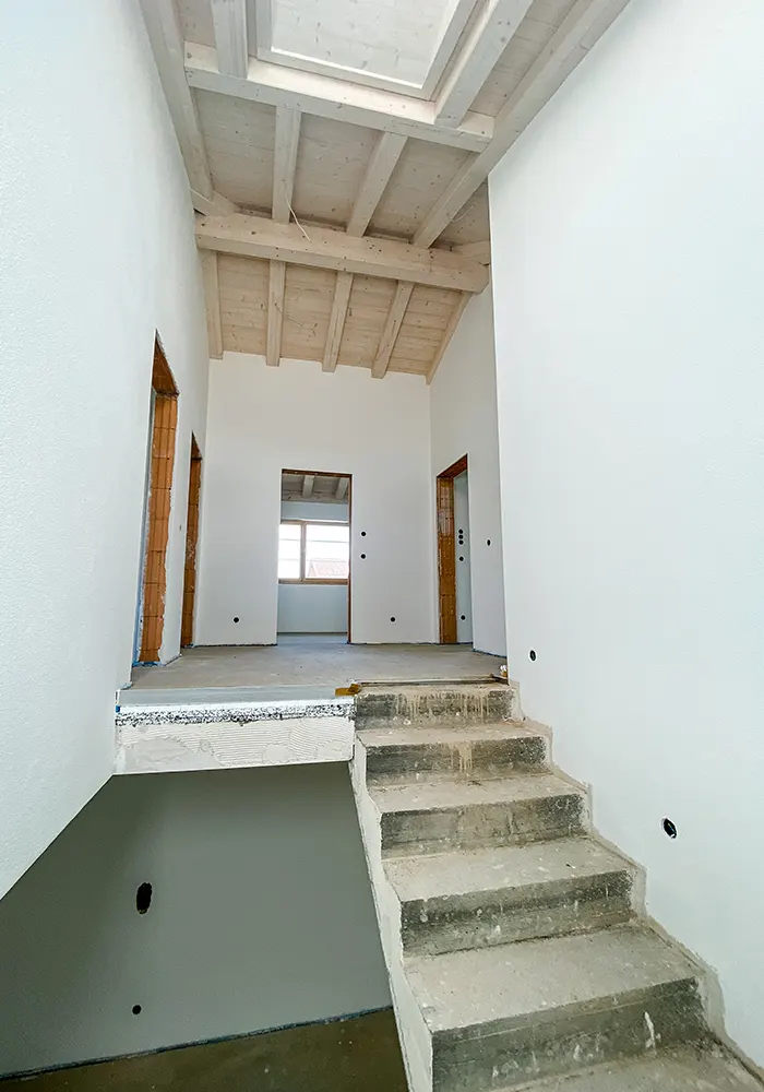 Rohbau eines Treppenhauses mit offener Holzdecke, weißen Wänden und mehreren Türöffnungen im Obergeschoss.