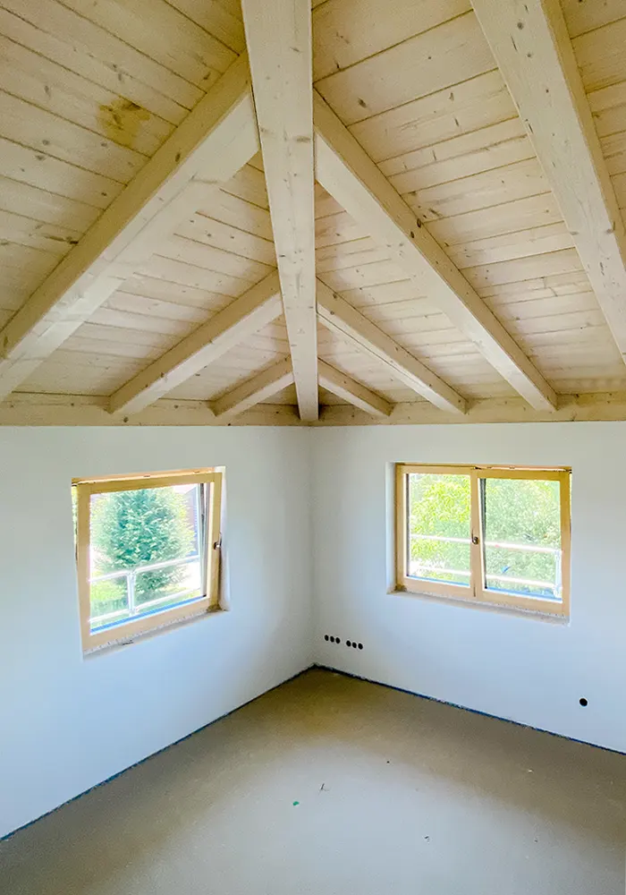 Heller leerer Raum mit zwei Fenstern und sichtbarem Holzbalken-Dachstuhl in naturbelassener Optik.
