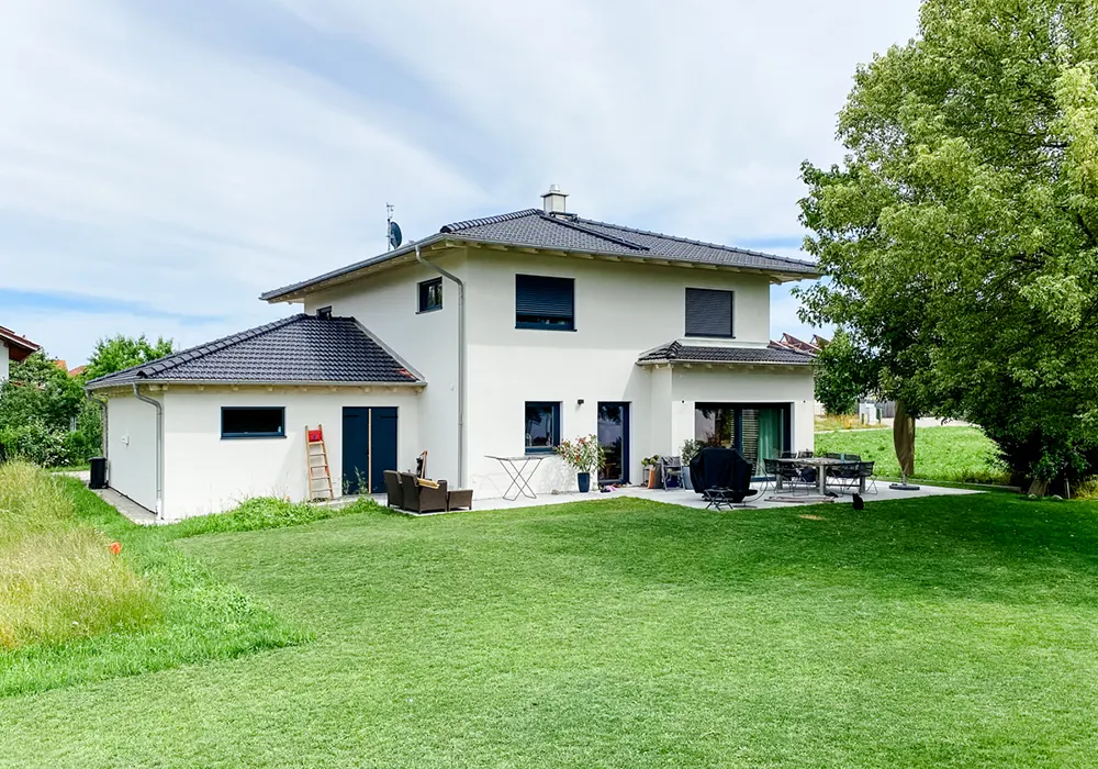 Modernes Einfamilienhaus mit großer Terrasse und gepflegtem Garten, umgeben von Bäumen und Wiese.