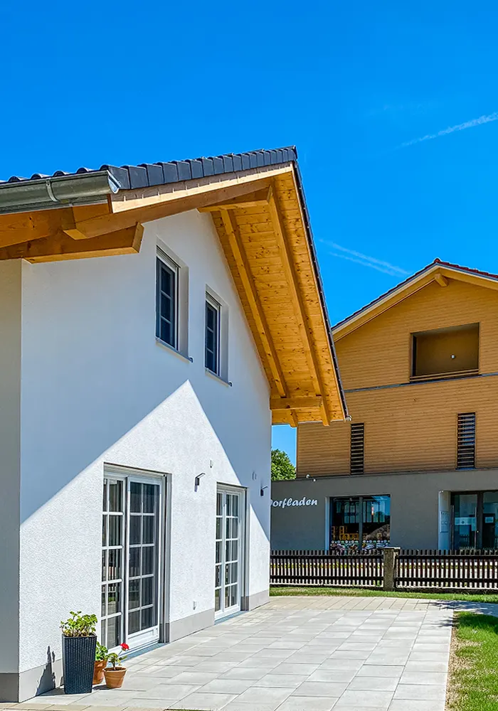 Nahaufnahme eines modernen Hauses mit weißer Fassade, Holzdach und großer Terrasse unter blauem Himmel.