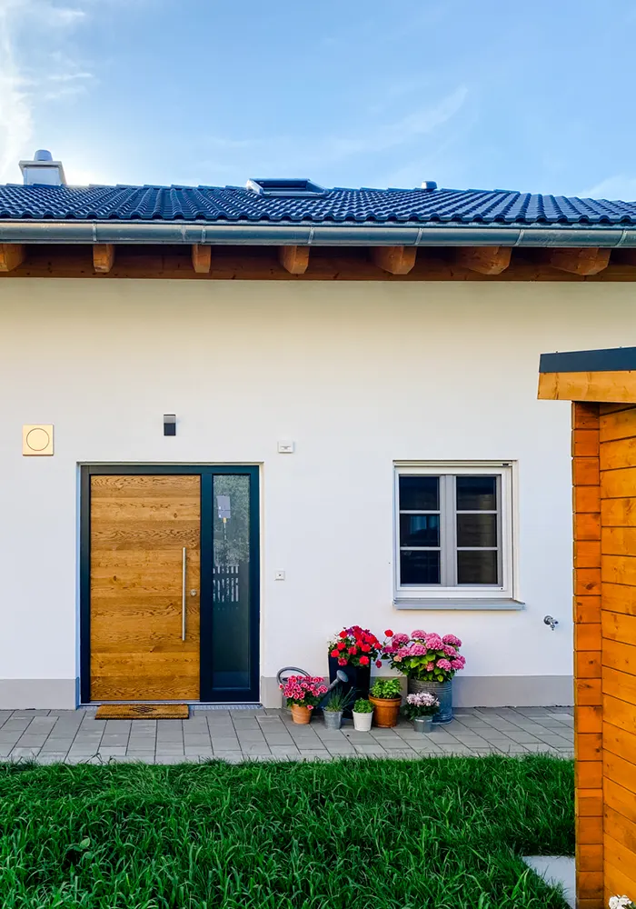 Eingangsbereich eines Hauses mit Holztür, seitlichem Fenster und bunten Blumentöpfen auf gepflasterter Terrasse.