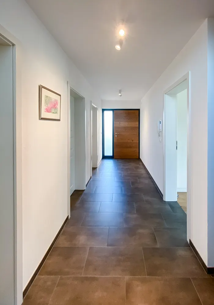 Heller Flur mit braunen Bodenfliesen, weißen Wänden und Holzeingangstür am Ende des Gangs.