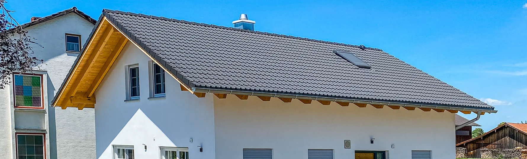 Modernes Haus mit Satteldach und Dachfenster, daneben ältere Gebäude unter blauem Himmel.