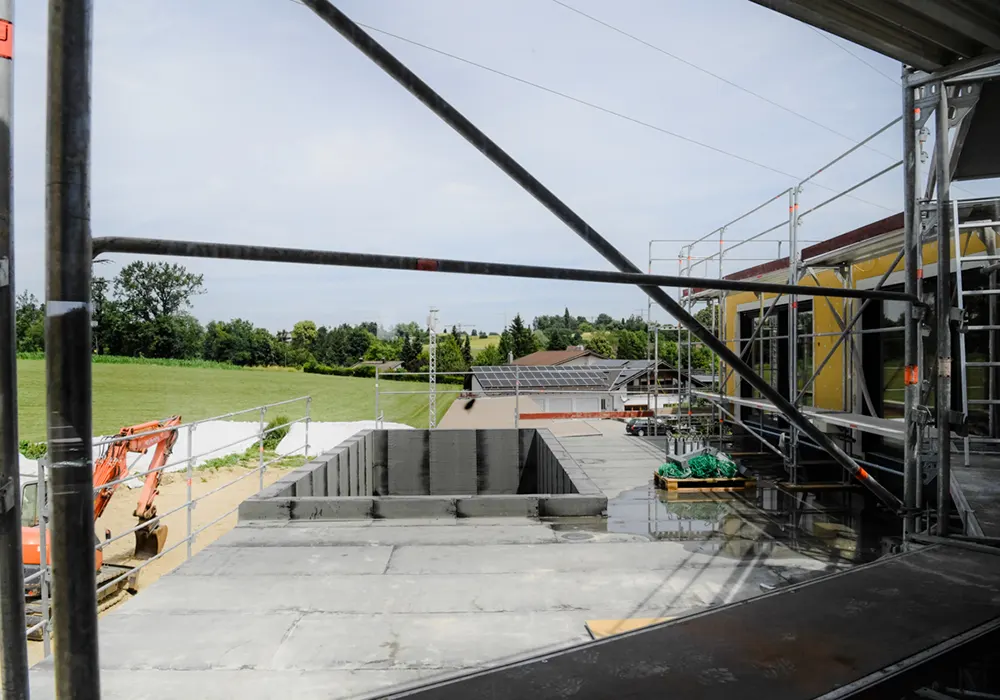 Blick von Gerüst auf Flachdach mit Baustelle, Öffnung in der Mitte und umliegender grüner Landschaft.