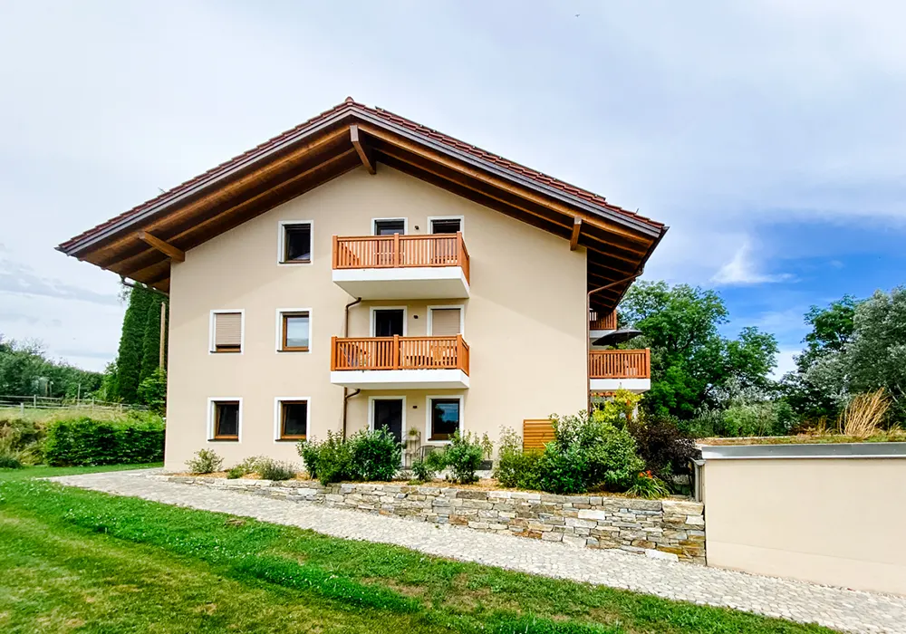 Beiges Mehrfamilienhaus mit roten Holzbalkonen, Gartenweg und Grünfläche im Vordergrund.